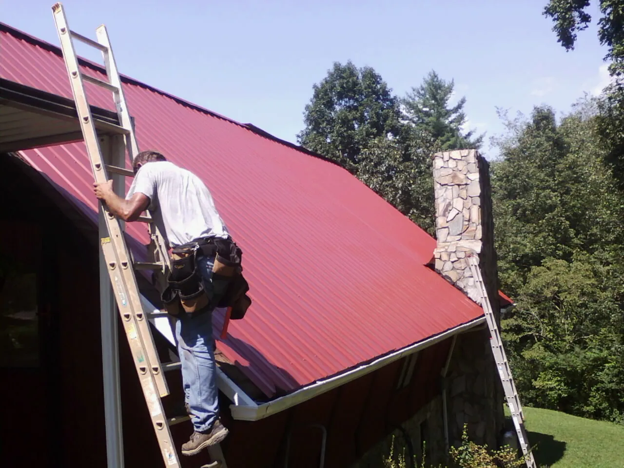 Roofer on a ladder beside a red metal roof and stone chimney — Soffit & Fascia Repair by Roofing Brothers in Valley Spring